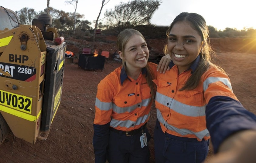 TWO WOMEN IN PILBARA WEARING PPE GEAR WITH BHP EQUIPMENT IN THE BACKGROUND