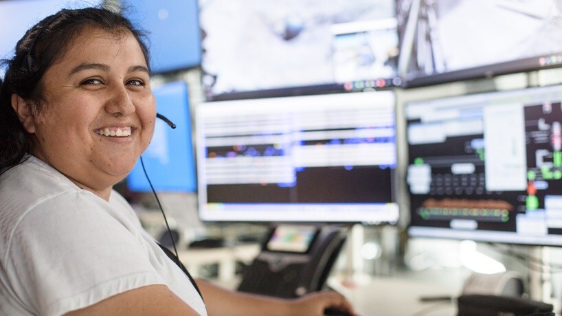 Smiling woman at desk with lots of computer monitors around her for remote controlled operations of a mine site