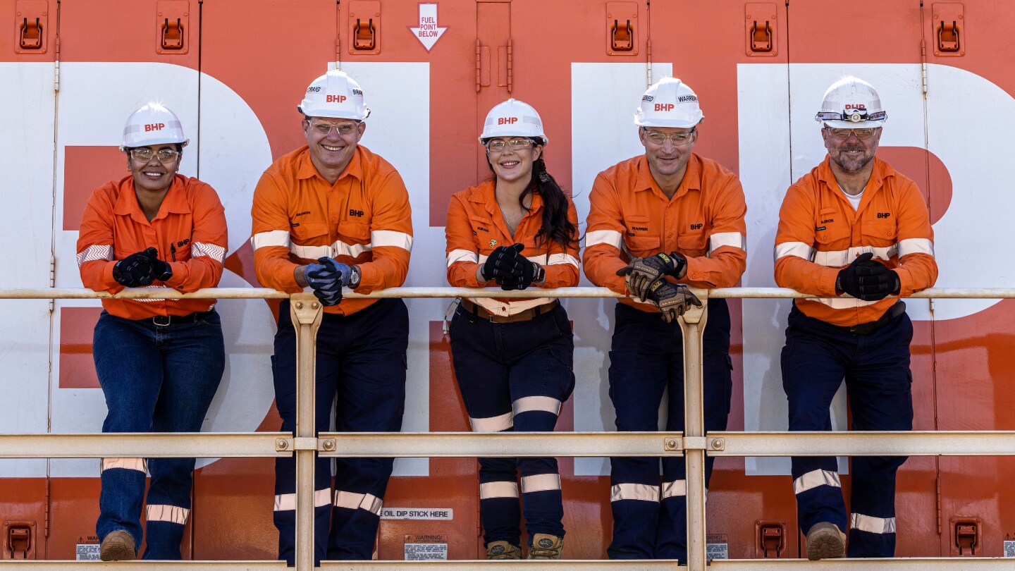 Five BHP men and women in orange shirts in front of BHP sign smiling wearing orange PPE