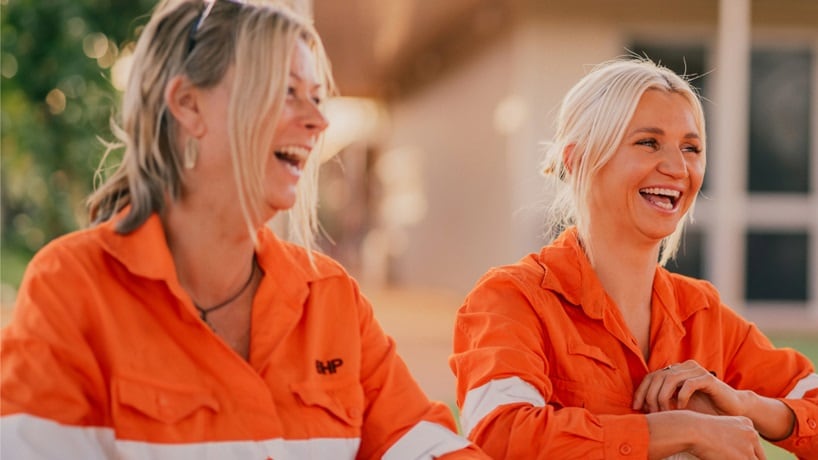 Two women wearing BHP orange shirts shown laughing at picnic tables as the sun sets 