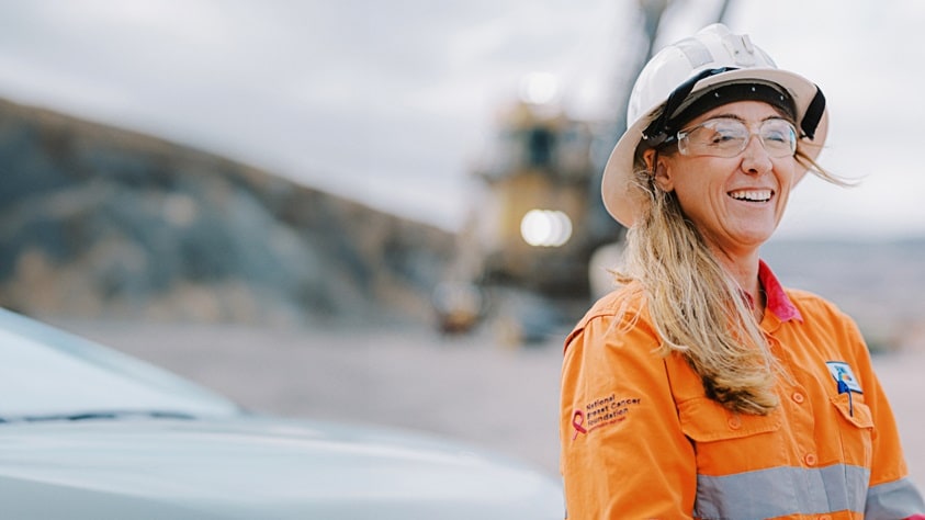 Woman in BHP orange shirt and hard hat laughing, shown on work site with equipment in the background