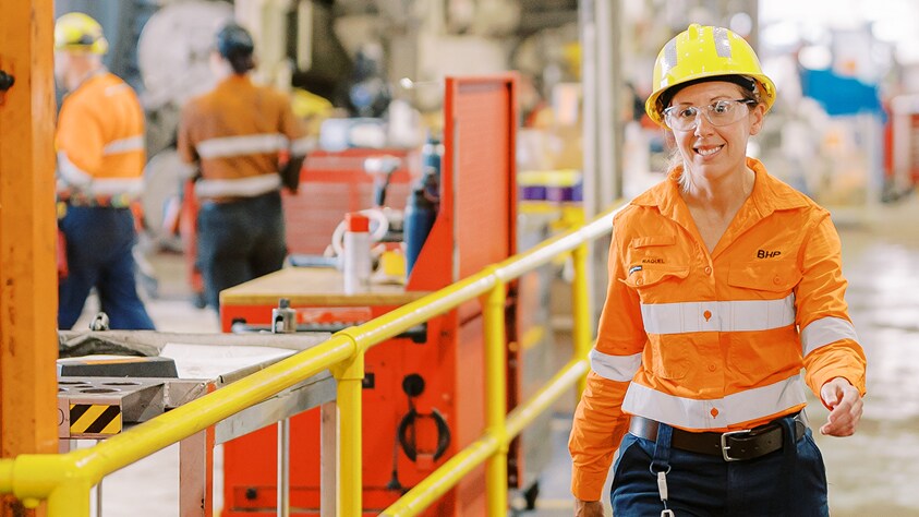 Raquel walking through maintenance workshop in BHP orange high vis shirt smiling with people shown in background