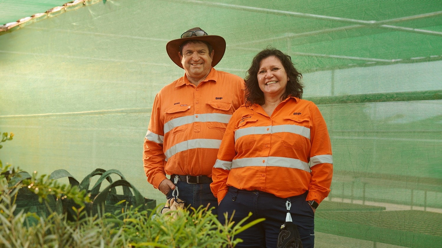 A man and woman, both smiling and in orange hi-vis shirts, stand inside a greenhouse.