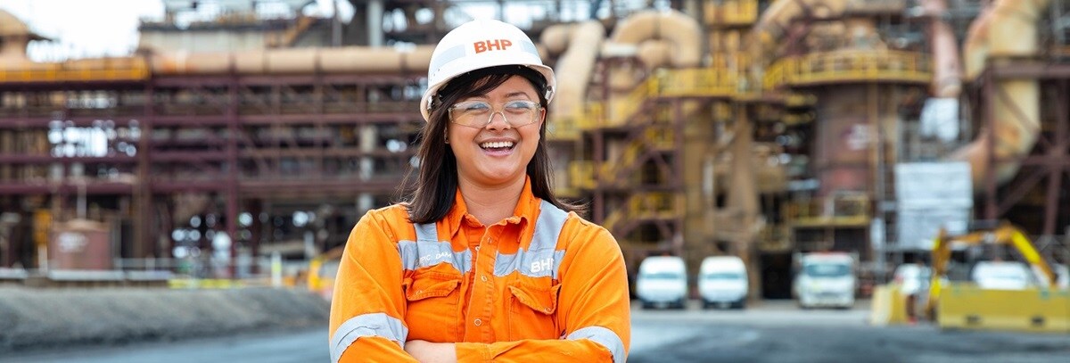 A woman, in hi-vis shirt and hard hat, stands with arms crossed and smiling. Olympic Dam infrastructure is in the background.