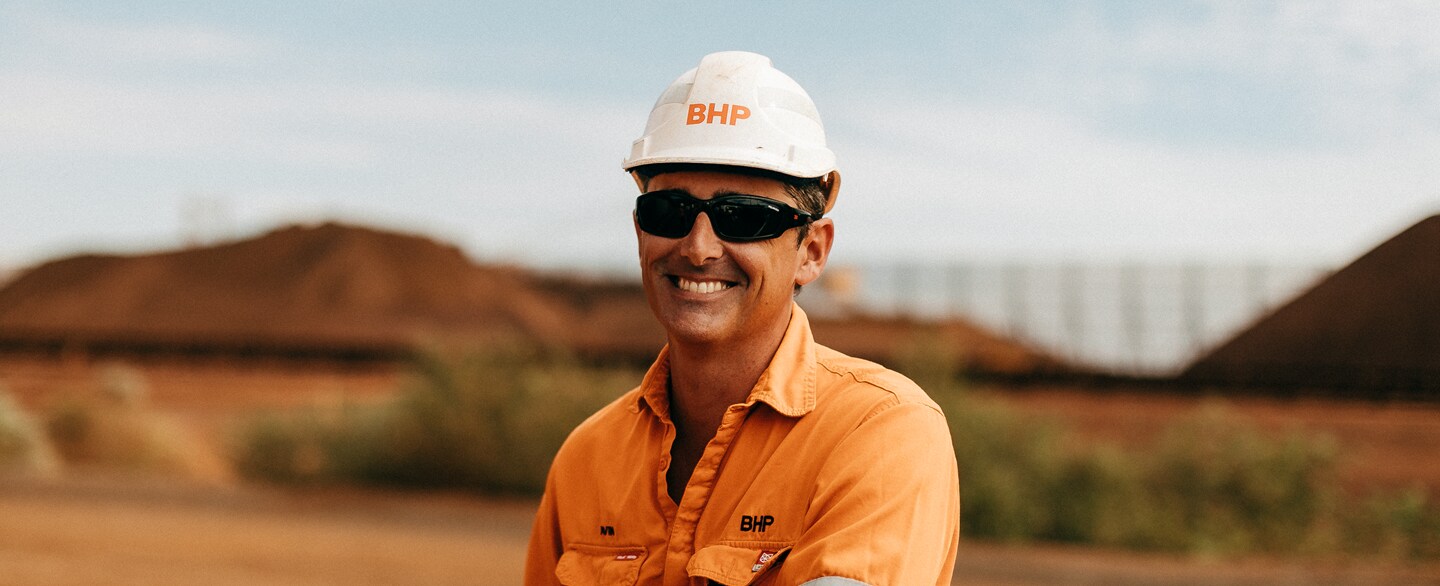 Man wearing orange shirt and white hard hat smiling at camera, with red dirt in background. 