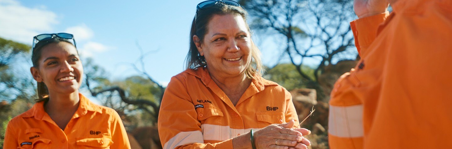 Two Indigenous women stand smiling in high-vis shirts. One is holding a piece of vegetation. 