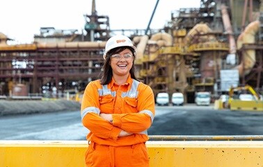 A woman, in hi-vis shirt and hard hat, stands with arms crossed and smiling. Olympic Dam infrastructure is in the background.