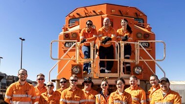 BHP team of men and women smiling in front of locomotive dressed in working PPE orange shirts