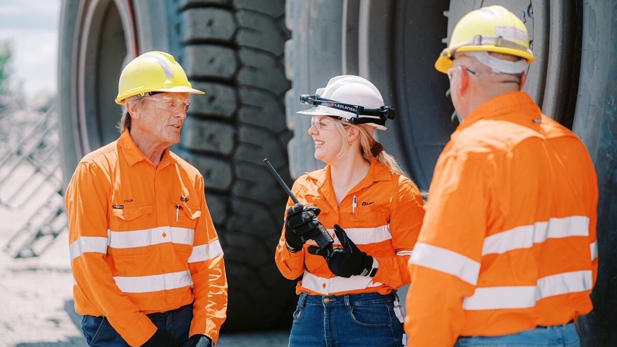 Three people in BHP orange PPE shown in front of large truck wheel smiling 