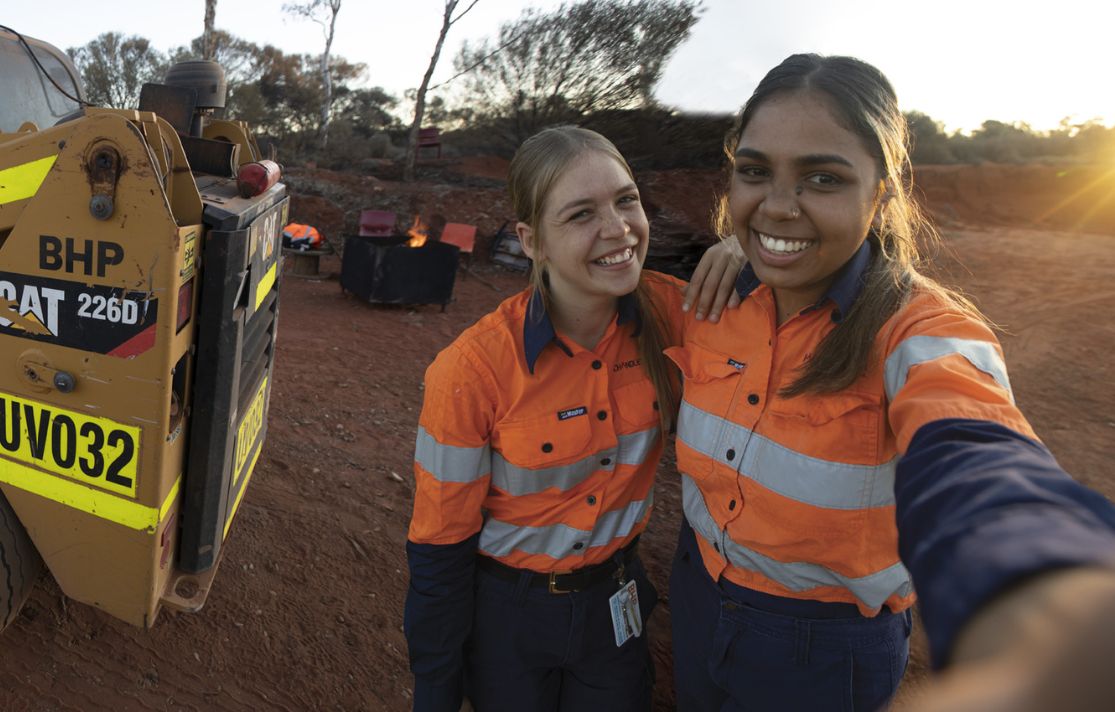 TWO WOMEN IN PILBARA WEARING PPE GEAR WITH BHP EQUIPMENT IN THE BACKGROUND