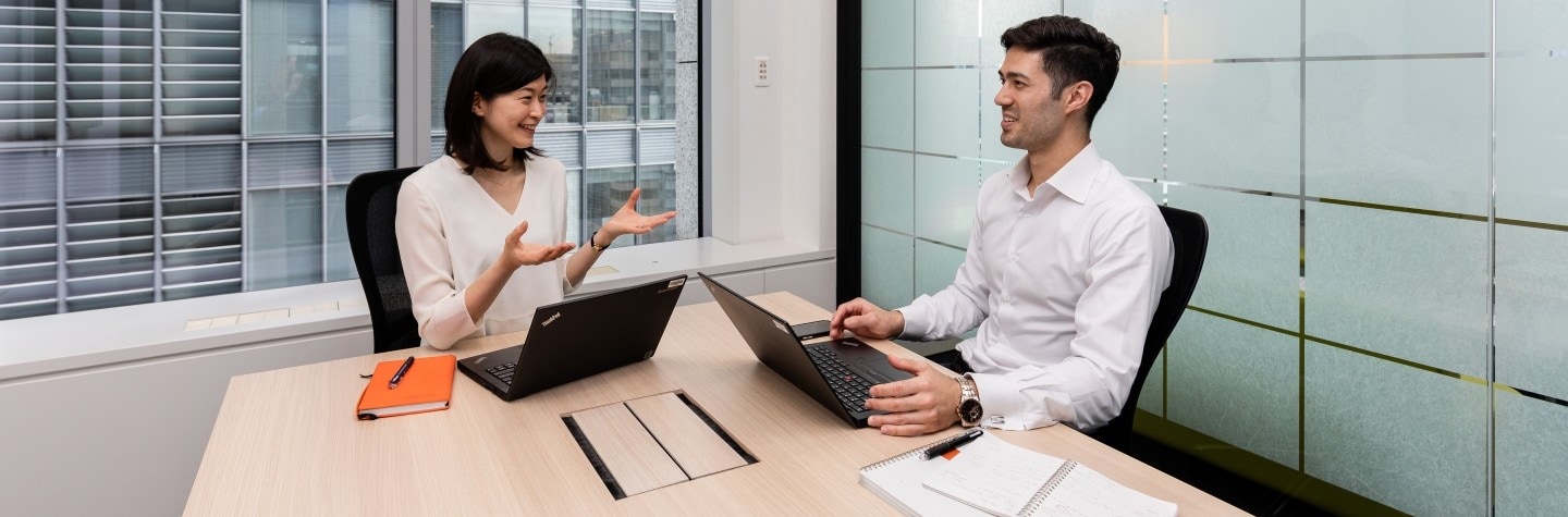 A woman and a man sit at table in an office in front of their laptops while talking
