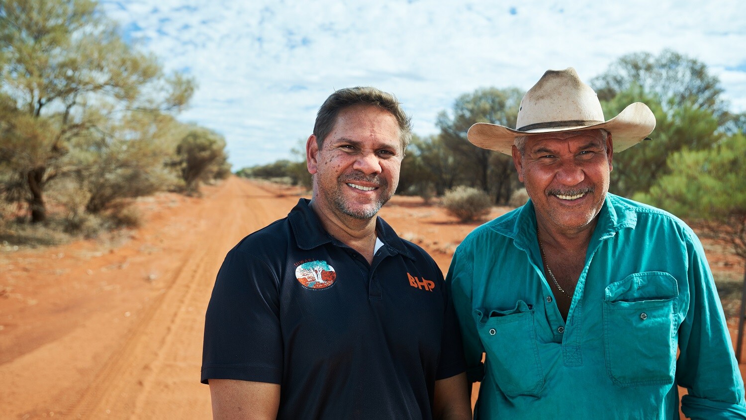 Two men stand smiling on a red dirt track. One is in a green shirt and akubra, the other is in a black BHP polo shirt. 
