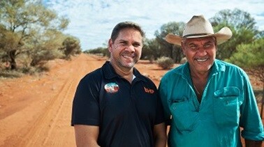 Two men, one in a black BHP shirt and one in a green shirt and akubra, stand on a red dirt road with green vegetation and blue sky behind them.