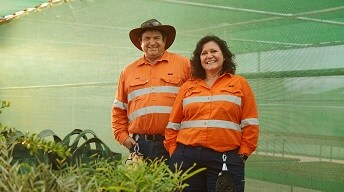Two people, a man and a woman, in high-vis shirts stand in a green house with some plants in front of them. They are similing. 