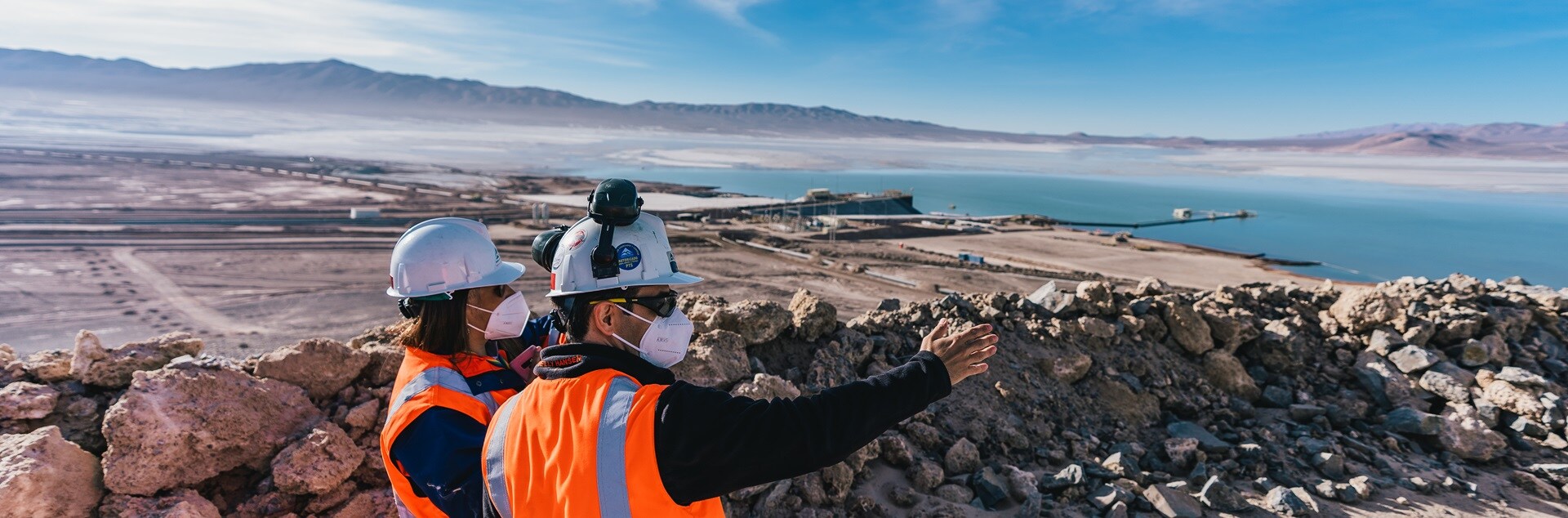 Two workers in high-visibility orange vests and personal protective equipment looking out over Escondida tailings storage facilities with mountains in the distance. 