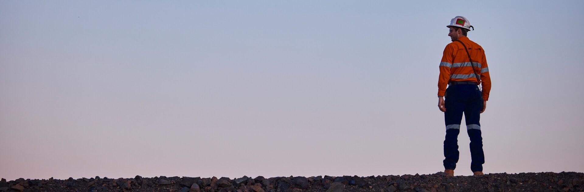 Frontline worker at Prominent Hill, South Australia, stands on a rocky ridge in high-visibility safety gear and hard hat, overlooking the mine site.